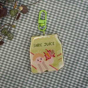 Puede incluir: Un llavero de plástico transparente con una ilustración de dibujos animados de un personaje sosteniendo una zanahoria. El llavero tiene el texto "SHIRE JUICE" y un clip verde.