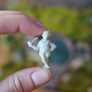 May include: A small, off-white figurine of a man in a period outfit, holding a pipe. The miniature figure is held between fingers against a blurred green and brown background. The figure is approximately 1 inch tall.