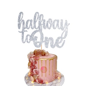 Puede incluir: Topper de pastel de purpurina plateada con el texto "halfway to one". El topper est&aacute; encima de un pastel rosa con glaseado dorado de goteo y decoraciones rosas y doradas.