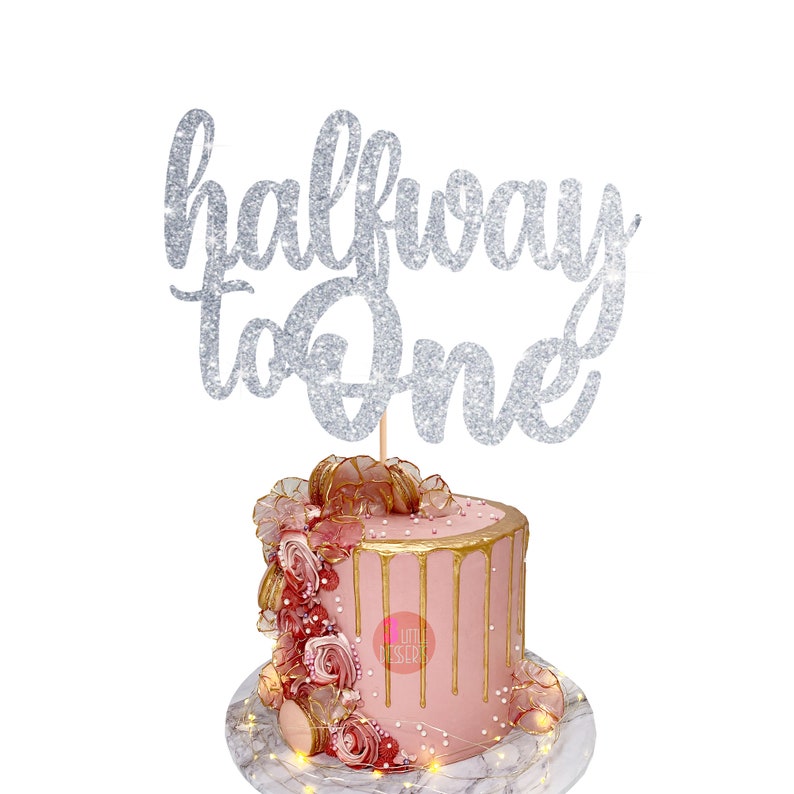 Puede incluir: Topper de pastel de purpurina plateada con el texto "halfway to one". El topper est&aacute; encima de un pastel rosa con glaseado dorado de goteo y decoraciones rosas y doradas.