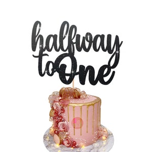Puede incluir: Topper de pastel de purpurina negra con el texto "halfway to one". El topper est&aacute; encima de un pastel rosa con glaseado dorado de goteo y decoraciones rosas y doradas.
