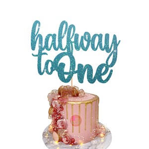 Puede incluir: Topper de pastel de purpurina turquesa con el texto "halfway to one". El topper est&aacute; encima de un pastel rosa con glaseado dorado de goteo y decoraciones rosas y doradas.