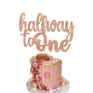 Puede incluir: Un pastel rosa con glaseado dorado y decoraciones de rosas coronado con un adorno de pastel de purpurina dorada que dice "halfway to one".