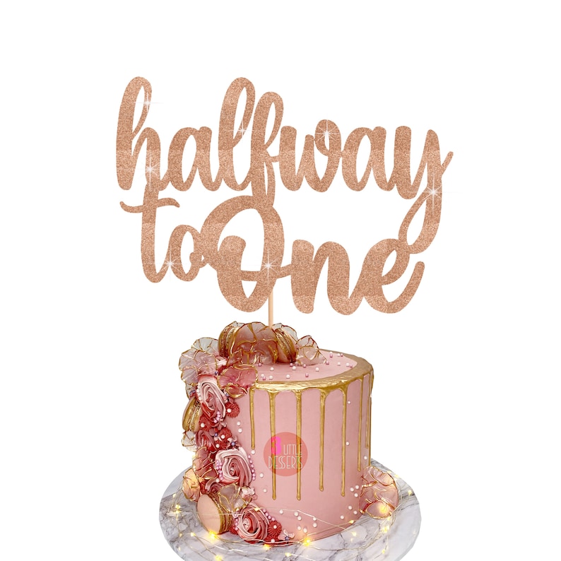 Puede incluir: Un pastel rosa con glaseado dorado y decoraciones de rosas coronado con un adorno de pastel de purpurina dorada que dice "halfway to one".