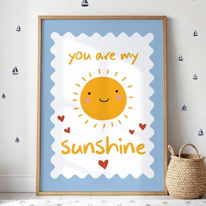 May include: A rectangular framed wall art print featuring a light blue background with a white scalloped border. In the centre is a yellow sun illustration with a smiling face and pink cheeks, surrounded by the text you are my Sunshine and red hearts.