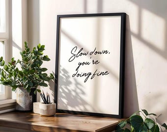 May include: A black framed wall art print featuring the black cursive text "Slow down, you're doing fine" on a cream background. The print is displayed upright on a wooden surface next to a potted green plant and a small container with pencils.