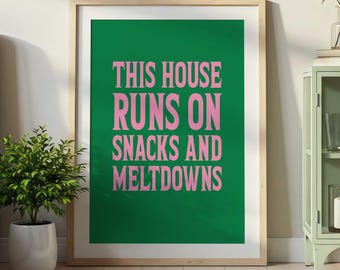 May include: A framed wall art print featuring a solid dark green background with the text "THIS HOUSE RUNS ON SNACKS AND MELTDOWNS" printed in a bold, light pink sans-serif font. The print is enclosed in a light-colored wooden frame.