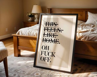May include: A framed rectangular wall art piece featuring a beige background with black text. The text reads: THIS FEELS. LIKE HOME. OH FUCK OFF. The first three lines have a horizontal black line striking through the center of the words.