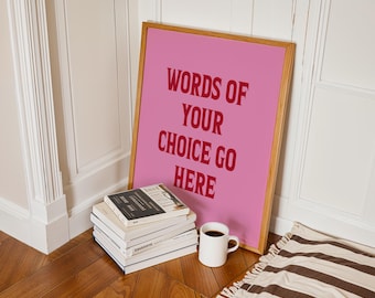 May include: A rectangular framed print with a pink background and bold red text that reads "WORDS OF YOUR CHOICE GO HERE". The frame is made of light-colored wood and is positioned upright against a white wall next to a stack of books.