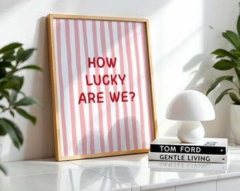 May include: A rectangular framed print featuring vertical pink and white stripes with the red text "HOW LUCKY ARE WE?" centered on the surface. The frame is made of light-colored wood and holds the striped paper artwork in a vertical orientation.