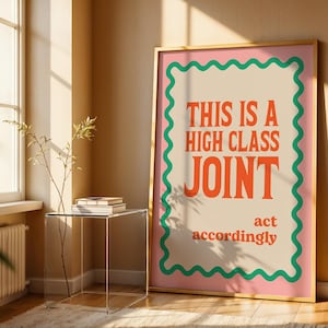 May include: A rectangular framed wall art print featuring a beige background with a pink border and a green wavy line. The text in orange reads: THIS IS A HIGH CLASS JOINT act accordingly. The print is displayed in a light-coloured wooden frame.