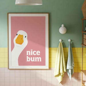 May include: A rectangular wall art print featuring a white goose with an orange beak on a solid pink background. The text "nice bum" is printed in white, bold, sans-serif letters to the right of the goose's neck within the frame.