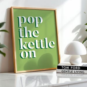 May include: A framed print with the text "pop the kettle on" in white on a green backdrop. A white mushroom lamp sits on two books, with a potted plant visible. The frame is wooden.