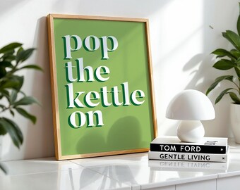 Peut inclure: Une affiche encadrée avec le texte "pop the kettle on" en blanc sur fond vert. Une lampe blanche en forme de champignon est posée sur deux livres, avec une plante en pot en arrière-plan.