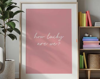 May include: A rectangular wall art print in a light wood frame featuring a dusty rose background with the white cursive text "how lucky are we?" centered on the print. The artwork is displayed in a clean, simple, and modern style.
