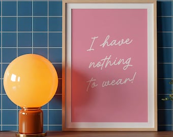 May include: A rectangular wall art print in a light wood frame featuring a pink background with the white cursive text I have nothing to wear! positioned next to a spherical orange glass lamp with a matching base on a wooden surface.
