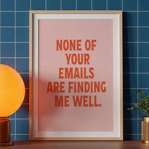May include: A rectangular wall art print in a light wood frame featuring a pale pink background with bold orange text that reads: NONE OF YOUR EMAILS ARE FINDING ME WELL. The print is centred and displays clear, sans-serif typography.