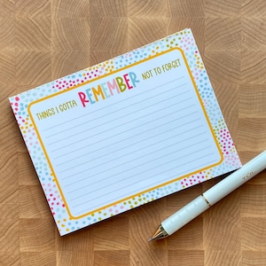 May include: A colorful notepad with the words "REMEMBER NOT TO FORGET" and "THINGS I GOTTA" printed on it. The notepad has a yellow border and is lined for writing. A white pen with gold accents rests on the side.