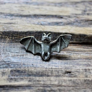 May include: A silver-coloured metal bat figurine with outstretched wings. The bat is in a flying position with its wings spread wide. The figurine is on a wooden surface.