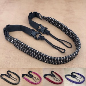 May include: A black and silver camera strap with a woven design and black plastic buckles. The strap has a loop at each end for attachment. Additional straps in gray, pink, red, and purple are shown below.