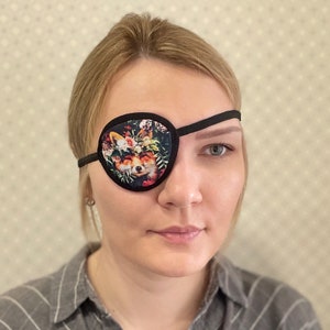 Op de afbeelding: Een zwart ooglapje met een bloemen vos ontwerp. De patch heeft een zwarte rand en band, met een kleurrijke afbeelding van een vos omringd door bloemen. Het ooglapje is ontworpen om één oog te bedekken.