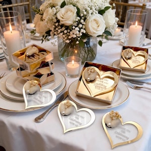 May include: Gold-rimmed heart-shaped acrylic favors with white floral accents and a pearl, featuring the text Thank You For Celebrating With Us. These items are displayed on white plates alongside gold gift boxes and lit candles on a table.