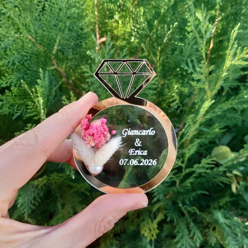 May include: A gold mirror ring with a diamond design and a small bouquet of dried flowers inside. The ring has the text "Giancarlo & Erica 07.06.2026" engraved on it.
