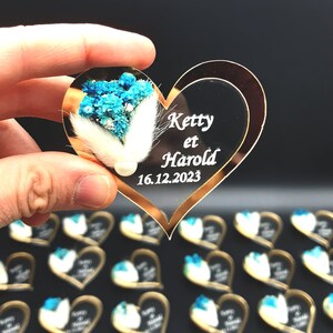 May include: A heart-shaped acrylic ornament with a gold-toned border, featuring white text that reads Ketty et Harold and 16.12.2023. The surface is decorated with a cluster of dried blue flowers, white fluffy bunny tail grass, and a small white pearl.