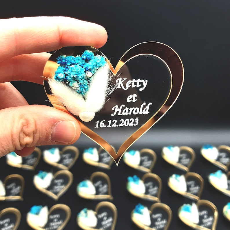 May include: A heart-shaped acrylic ornament with a gold-toned border, featuring white text that reads Ketty et Harold and 16.12.2023. The surface is decorated with a cluster of dried blue flowers, white fluffy bunny tail grass, and a small white pearl.