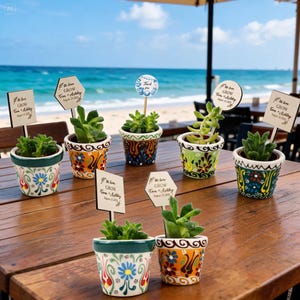 Puede incluir: Siete pequeñas plantas suculentas en macetas de cerámica pintadas a mano con coloridos motivos florales. Cada maceta tiene una estaca de madera con un letrero que dice "Let love GROW Tom + Ashley August 22, 2022" o una etiqueta "Thank you".