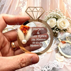 May include: A gold-toned acrylic ring-shaped ornament featuring a diamond cutout at the top. The surface is engraved with the text Meryem & Scott Kerem, S&ouml;z Hatırası, 01-03-2024, İlkbaharın İlk G&uuml;n&uuml;, accented with a small orange dried flower and a white pearl.