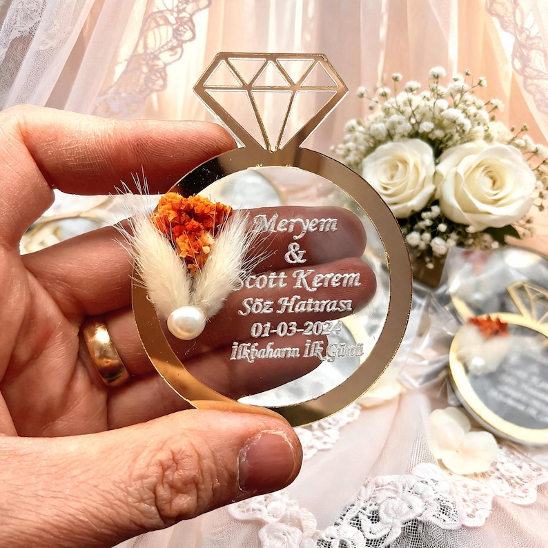 May include: A gold-toned acrylic ring-shaped ornament featuring a diamond cutout at the top. The surface is engraved with the text Meryem & Scott Kerem, S&ouml;z Hatırası, 01-03-2024, İlkbaharın İlk G&uuml;n&uuml;, accented with a small orange dried flower and a white pearl.
