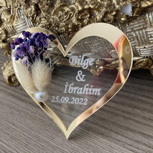 May include: A gold heart-shaped mirror with a white inscription that reads "Bilge & Ibrahim 25.09.2022". The mirror is decorated with dried flowers and a pearl.