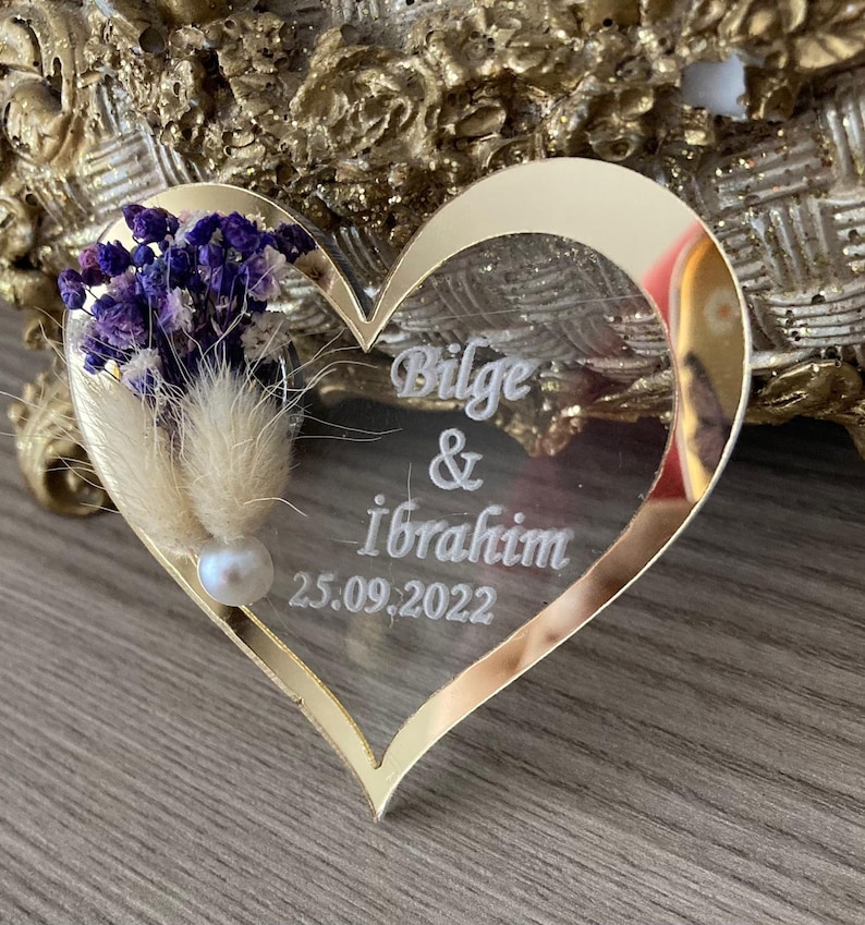 May include: A gold heart-shaped mirror with a white inscription that reads "Bilge & Ibrahim 25.09.2022". The mirror is decorated with dried flowers and a pearl.