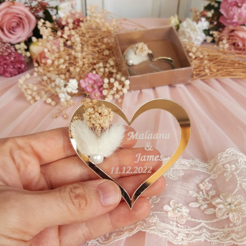 May include: A heart-shaped gold mirror ornament featuring white dried flowers, a small white pearl, and the engraved text Malaana & James 11.12.2022. The reflective heart is held in place, showcasing its smooth metallic finish and floral detail.