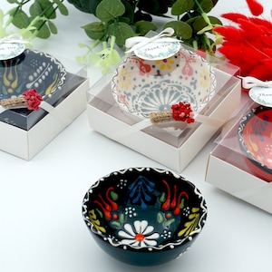 Op de afbeelding: Vier kleine keramische kommen met kleurrijke bloemenpatronen, gepresenteerd in transparante geschenkdozen met witte deksels en wit satijnen lint. Elke doos heeft een klein label met de tekst "Bedankt".