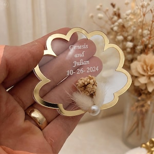 May include: A cloud-shaped clear acrylic ornament with a gold-colored reflective border. The center features white engraved text reading Genesis and Julian 10-26-2024. A small cluster of dried beige flowers and a single white pearl are attached to the front.
