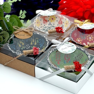 May include: Four small ceramic bowls in clear boxes with ribbons and tags reading Thank You. The bowls feature colorful floral patterns in black, green, red, and cream. Each box is decorated with a small bundle of dried red flowers and twine.