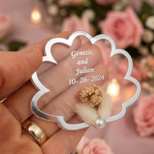 May include: A cloud-shaped clear acrylic ornament with a silver-toned border. The center features white engraved text reading Genesis and Julian 10-26-2024. A small cluster of dried beige flowers and a single white pearl bead are attached to the bottom.