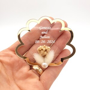 May include: A scalloped, gold-rimmed clear acrylic ornament featuring the engraved text Genesis and Julian 10-26-2024. The center is decorated with a small cluster of dried beige flowers, fluffy white grass, and a single white pearl bead.
