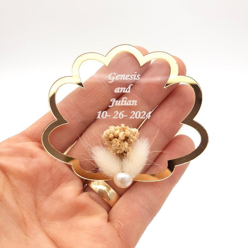 May include: A scalloped, gold-rimmed clear acrylic ornament featuring the engraved text Genesis and Julian 10-26-2024. The center is decorated with a small cluster of dried beige flowers, fluffy white grass, and a single white pearl bead.