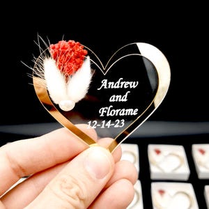 May include: A heart-shaped black acrylic ornament featuring the white engraved text Andrew and Florame 12-14-23. The ornament is decorated with a cluster of dried red flowers and two white fluffy bunny tail grass stems with a small white pearl bead.
