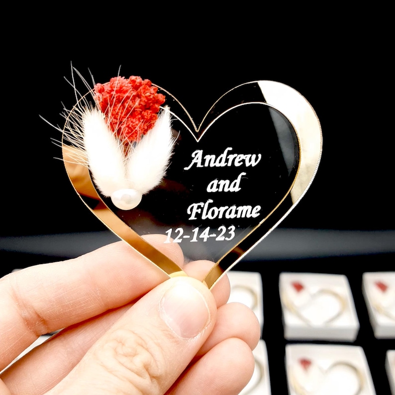 May include: A heart-shaped black acrylic ornament featuring the white engraved text Andrew and Florame 12-14-23. The ornament is decorated with a cluster of dried red flowers and two white fluffy bunny tail grass stems with a small white pearl bead.