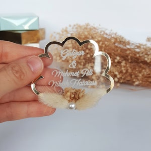 May include: Gold-rimmed clear acrylic wedding favor with the text "G&uuml;lizar & Mehmet Ali Nikah Hatırası" and a small bouquet of dried flowers.