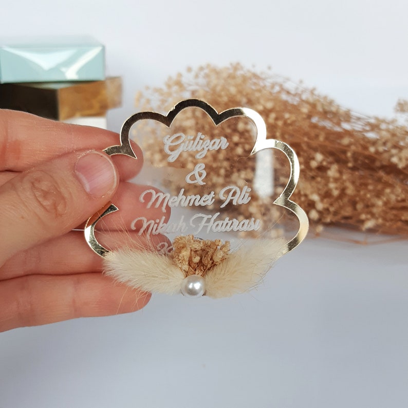 May include: Gold-rimmed clear acrylic wedding favor with the text "G&uuml;lizar & Mehmet Ali Nikah Hatırası" and a small bouquet of dried flowers.