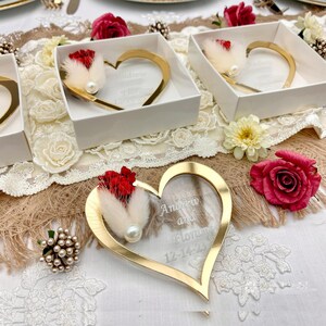 May include: A heart-shaped acrylic ornament with a gold-toned metallic border, featuring the engraved text Andrew and Floriane 12-14-23. The ornament is decorated with a cluster of red dried flowers, fluffy white bunny tail grass, and a white pearl.