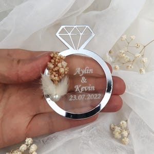 May include: A circular silver-toned acrylic ornament shaped like a diamond ring. The center is transparent and engraved with the text Aylin & Kevin 23.07.2022. It is decorated with small beige dried flowers and a single white pearl-like bead.