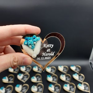 May include: A clear heart-shaped acrylic ornament with a gold border. The ornament features a small bouquet of blue flowers and a white feather. The text "Ketty et Harold 16.12.2023" is engraved on the ornament.