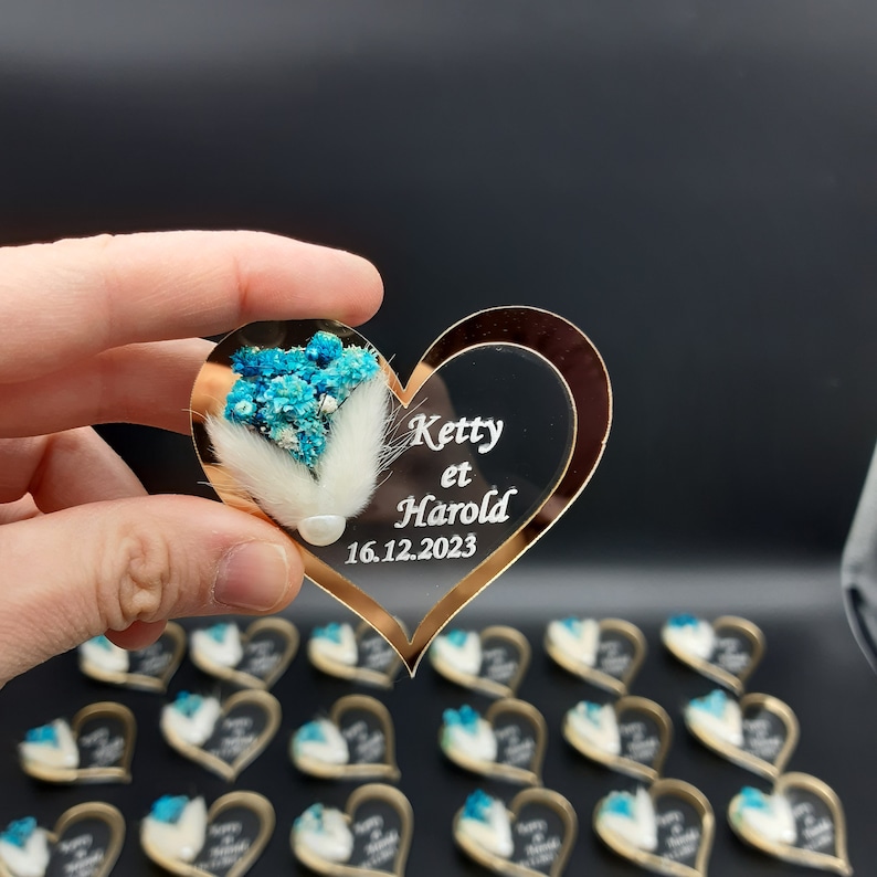 May include: A clear heart-shaped acrylic ornament with a gold border. The ornament features a small bouquet of blue flowers and a white feather. The text "Ketty et Harold 16.12.2023" is engraved on the ornament.
