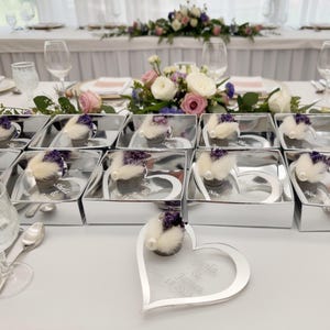 May include: A collection of silver heart-shaped acrylic frames, each featuring a small cluster of dried purple flowers and white pampas grass with a pearl accent. The frames are engraved with the text Sheila & James Nov 18 2023.